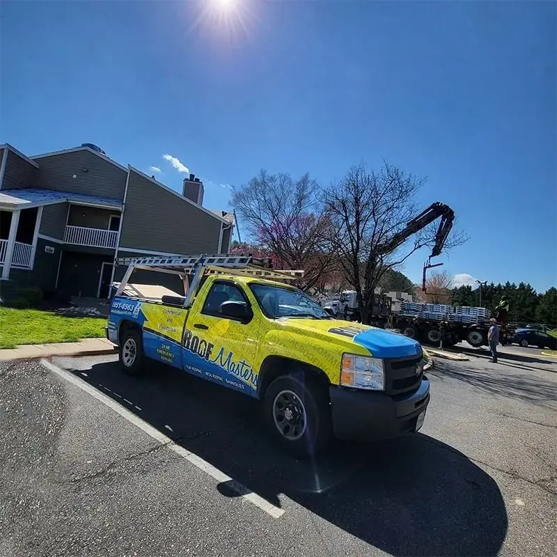The yellow and blue Roof Masters truck parked at an apartment re-roof while a crane lifts bundles of shingles
